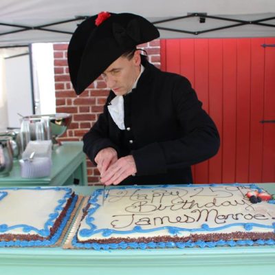 Man dressed as James Monroe, hat and all cutting a big sheet cake saying "Happy 259th Birthday James Monroe!"